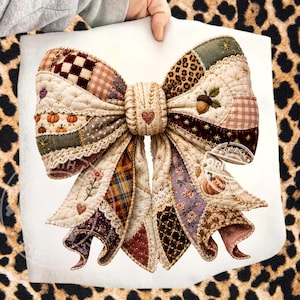 May include: A large decorative bow made of quilted fabric patches in various patterns including plaid, gingham, leopard print, and floral motifs. The bow features embroidered details like pumpkins, an acorn, and hearts, with cream lace trim edges.
