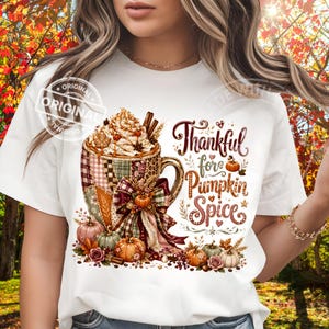 May include: A white t-shirt featuring a graphic of a mug filled with whipped cream and cinnamon sticks. The mug has a patchwork pattern and a bow. Text reads Thankful for Pumpkin Spice. Surrounding the mug are small pumpkins and autumn leaves.