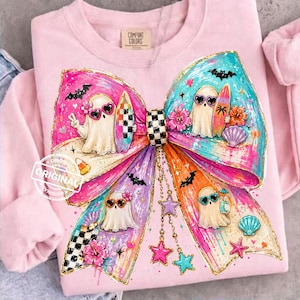 May include: Pale pink sweatshirt with a vibrant bow illustration. The bow design includes ghost characters with sunglasses, surfboards, and decorative elements like bats and flowers. The word "ORIGINAL" is printed on the sweatshirt.