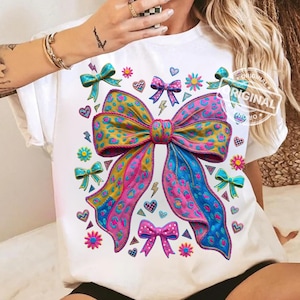 Puede incluir: Camiseta blanca con un gran diseño de lazo colorido con estampado de leopardo, cintas rosas y azules. La camiseta está decorada con lazos más pequeños, corazones, flores y rayos. La palabra "ORIGINAL" está impresa en la camiseta.