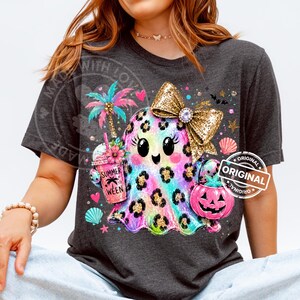 May include: A dark gray t-shirt featuring a colorful, leopard-print ghost graphic. The ghost wears a large gold glitter bow, holds a pink drink cup labeled SUMMER WEEN with a palm tree, and carries a pink jack-o'-lantern bucket with a starfish.
