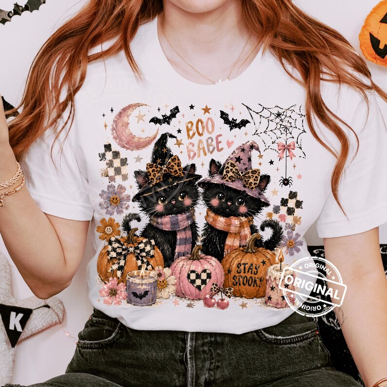 May include: White t-shirt featuring a Halloween-themed graphic. The design includes two black cats wearing witch hats and scarves, pumpkins, and the text "Boo Babe" and "Stay Spooky". The design incorporates floral and celestial elements.