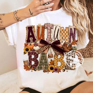 May include: A white t-shirt featuring a graphic design with the text AUTUMN BABE. The letters are filled with various patterns including plaid, leopard print, and polka dots. The design includes pumpkins, sunflowers, a dark red bow, and boots.