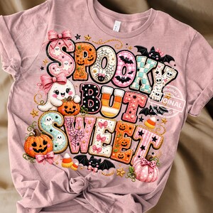 May include: A dusty rose t-shirt featuring a graphic print with the text "SPOOKY BUT SWEET". The design includes cartoon ghosts, jack-o'-lanterns, bats, candy corn, spider webs, and pink bows, all rendered in a colorful, stitched-style pattern.