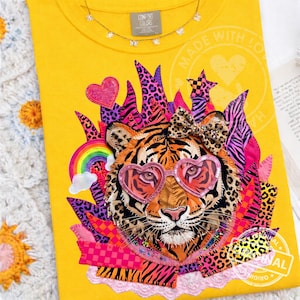 Può includere: T-shirt giallo con un design colorato a forma di faccia di tigre. La tigre indossa occhiali da sole a forma di cuore e un fiocco. Il design include un arcobaleno, stelle e la scritta "MADE WITH LOVE". La maglietta ha la scritta "COMFORT COLORS" sull'etichetta.