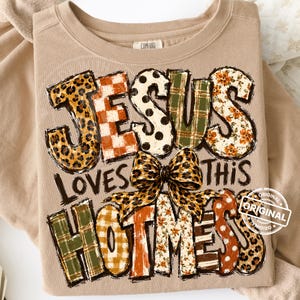 May include: A tan crewneck sweatshirt featuring the text JESUS LOVES THIS HOTMESS in large, multi-patterned block letters. The design includes leopard print, orange and white checkered, polka dot, green plaid, and floral patterns with a leopard print bow.