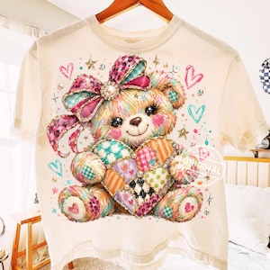 May include: A cream-colored t-shirt featuring a graphic of a teddy bear with a large pink leopard-print bow on its head. The bear holds a multi-colored patchwork heart. The design includes small stars, hearts, and circular accents in pink, blue, and orange.