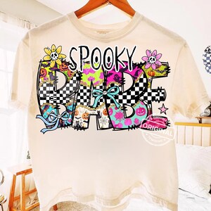 Op de afbeelding: Crèmekleurig T-shirt met de tekst "SPOOKY BABE" in een kleurrijk patroon. Het ontwerp bevat zwart-witte dambordpatronen, bloemen met schedels en Halloween-thema graphics zoals pompoenen en vleermuizen.