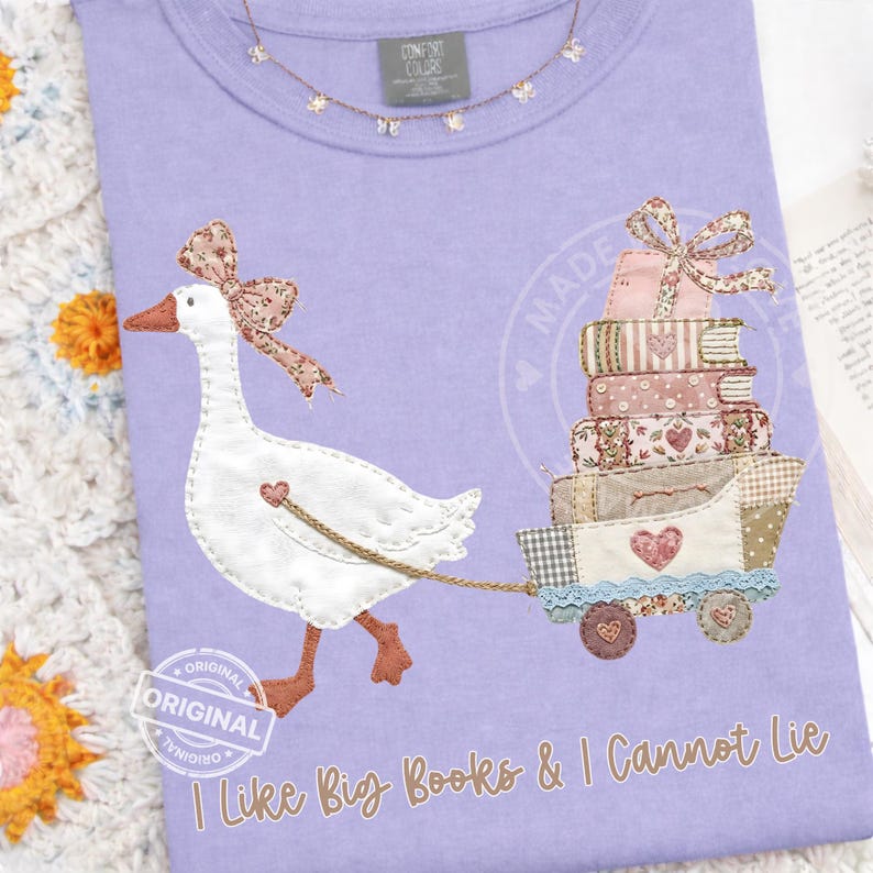 Puede incluir: Camiseta lila Comfort Colors con un ganso bordado tirando de un carro de libros. El ganso lleva un lazo y tira de un carro con un dise&ntilde;o de coraz&oacute;n. El texto "I Like Big Books & I Cannot Lie" est&aacute; impreso en la parte inferior.