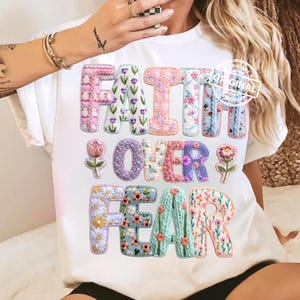 May include: White t-shirt featuring the embroidered phrase "FAITH OVER FEAR". The letters are decorated with floral patterns in various colours. The design includes flower illustrations, creating a vibrant graphic.