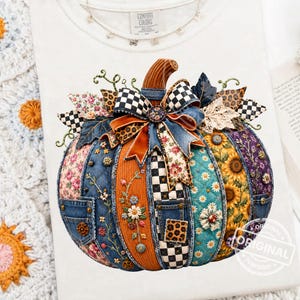 May include: A white t-shirt featuring a large, central graphic of a pumpkin constructed from various fabric patterns including denim, floral prints, leopard print, sunflowers, and black-and-white checkered squares, topped with a decorative bow.