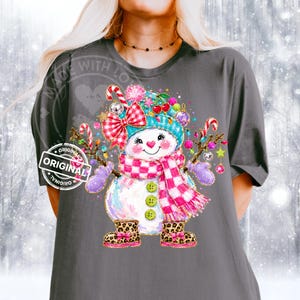 May include: A dark gray t-shirt featuring a graphic of a snowman wearing a teal hat, a pink and white checkered scarf, and leopard print boots. The snowman is decorated with a red and white bow, cherries, and candy canes with colorful ornaments.