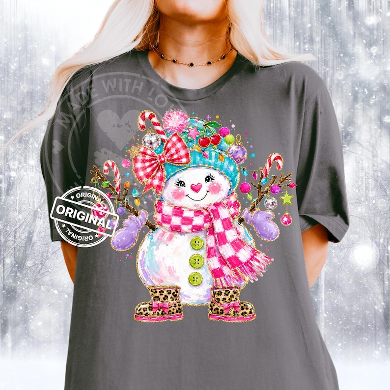 May include: A dark gray t-shirt featuring a graphic of a snowman wearing a teal hat, a pink and white checkered scarf, and leopard print boots. The snowman is decorated with a red and white bow, cherries, and candy canes with colorful ornaments.
