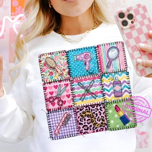 May include: White sweatshirt featuring a patchwork design of beauty-related icons. The patches include a hairbrush, hairdryer, mirror, scissors, bobby pins, hairspray, comb, leopard print, and lipstick. The design is stitched with black thread.