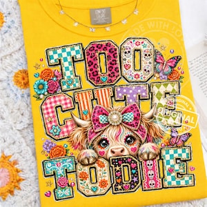 Könnte beinhalten: Ein leuchtend gelbes T-Shirt mit einer Grafik eines flauschigen Hochlandrindes, das eine rosa Schleife trägt. Der Text "TOO CUTE TO DIE" ist in großen, bunten, gemusterten Blockbuchstaben dargestellt, die mit Blumen, Schmetterlingen und Totenköpfen verziert sind.