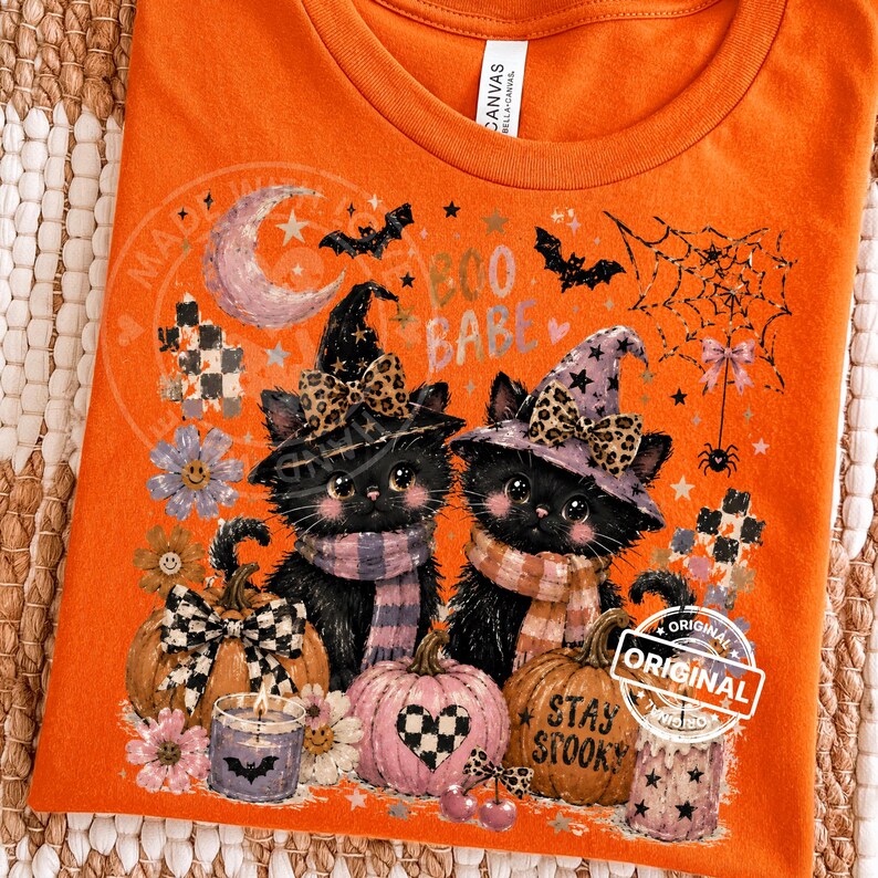 May include: Orange t-shirt with a Halloween-themed graphic. Two black cats wearing witch hats and scarves are featured, along with pumpkins, bats, and the text "Boo Babe" and "Stay Spooky".