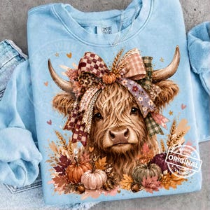 May include: A light blue crewneck sweatshirt featuring a graphic of a Highland cow with long horns, wearing a large multi-patterned bow. The design includes autumn-themed elements like pumpkins, wheat stalks, and fall leaves in orange and brown.