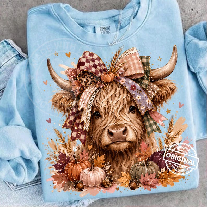 May include: A light blue crewneck sweatshirt featuring a graphic of a Highland cow with long horns, wearing a large multi-patterned bow. The design includes autumn-themed elements like pumpkins, wheat stalks, and fall leaves in orange and brown.