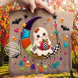 May include: A beige fabric square featuring a decorative applique of a white ghost holding a checkered pumpkin, sitting on a crescent moon made of colorful patterned patches. The design includes hanging beaded tassels, bats, hearts, and candy corn.