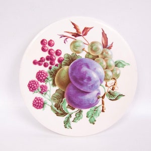 Pode incluir: Uma base para copos redonda de cerâmica branca com uma ilustração colorida de frutas. O design inclui ameixas roxas, uvas verdes, groselhas vermelhas e framboesas rosa, com folhas verdes e caules marrons. A base é um item decorativo.