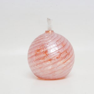 May include: A pink glass oil lamp with a spiral design. The lamp has a round body and a clear wick. The glass has a pearlescent sheen, and the lamp is set against a white background.