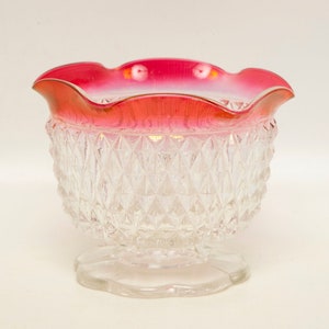 May include: A clear glass bowl with a diamond pattern and a ruffled, red rim. The bowl sits on a clear, scalloped base. The rim has a gold accent.