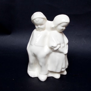 Könnte beinhalten: Eine weiße Keramikfigur, die zwei Figuren darstellt, einen Jungen und ein Mädchen, die eng beieinander stehen. Der Junge trägt einen Hut und eine Latzhose, während das Mädchen ein Kleid trägt. Die Figuren blicken nach unten.