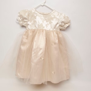 May include: A cream-colored baby dress with a floral pattern on the bodice and short, puffed sleeves. The skirt is a soft, flowing material with a tulle overlay. Small, decorative bows are sewn onto the skirt.