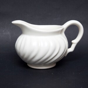 May include: A white ceramic creamer featuring a rounded, bulbous body with a distinct swirling, ribbed texture. The vessel includes a curved pouring spout on one side and a matching arched handle on the opposite side, set on a small circular base.