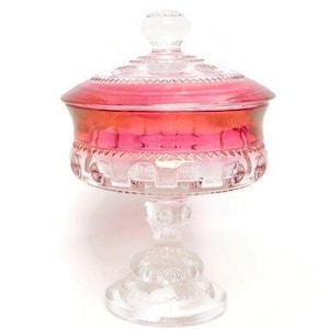 Pode incluir: Uma taça de vidro transparente com base de pedestal e tampa removível. A parte superior da taça e a tampa apresentam um gradiente rosa vibrante. O vidro é decorado com um padrão texturizado e em relevo por toda a sua superfície externa.
