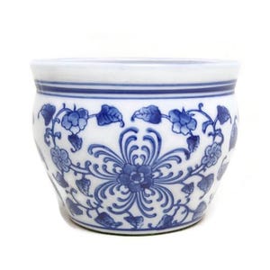 May include: A small, round, white ceramic planter with a blue floral design. The planter has a repeating pattern of flowers and leaves around the exterior. The top rim has a simple blue stripe. Suitable for small plants or succulents.