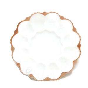 Peut inclure: Un plat blanc à bord festonné avec un bord de couleur cuivre. Le plat a une forme circulaire avec une série d'indentations arrondies sur le bord. L'intérieur est lisse et blanc, contrastant avec le bord métallique.