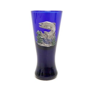 May include: A cobalt blue shot glass with a silver-colored fish and forest scene. The glass has a flared shape and a dark base. The fish is leaping out of the water, with trees in the background.
