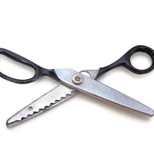 May include: A pair of dressmaking shears with black handles and silver blades. One blade has a zigzag edge, and the other is straight. The scissors are open, displaying the cutting edges. The background is plain white.
