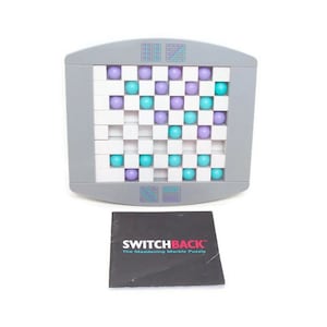May include: A gray plastic puzzle board with a grid of white squares containing purple and teal marbles, accompanied by a black instruction booklet labeled SWITCHBACK The Maddening Marble Puzzle in white and pink text on the front cover.