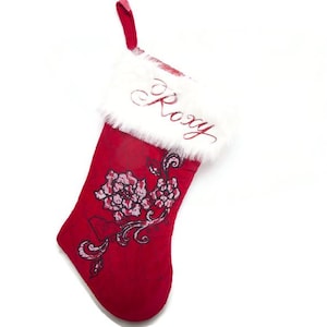 May include: Red Christmas stocking with white faux fur trim and a red loop for hanging. The stocking features a floral design and the name "Roxy" embroidered in red script. A festive holiday decoration.