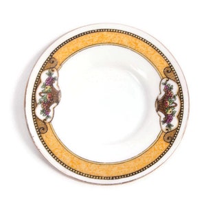 May include: Small, white ceramic plate with a decorative orange border and a fruit basket design. The border is accented with small black dots. The fruit basket design includes grapes, apples, and other fruits.