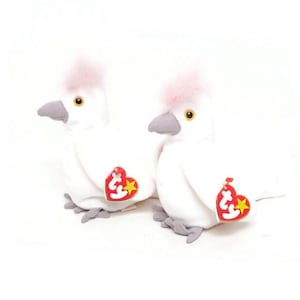 May include: Two white plush cockatoo toys with pink crests, gray beaks and feet, and yellow eyes. Each bird has a red heart-shaped tag with a yellow star. The toys are on a white background.