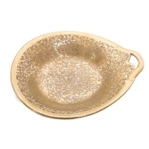 May include: A gold-colored, speckled ceramic serving dish with a handle. The dish has a teardrop shape with a textured interior and a smooth, light-colored rim. The handle has a decorative cutout.