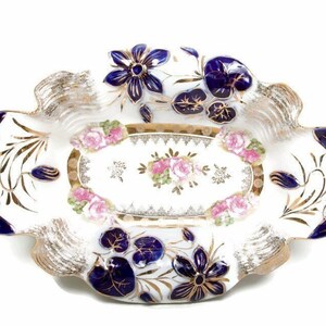 May include: White porcelain serving dish with a scalloped edge, featuring gold accents and floral designs. The dish has a central oval with pink roses and a border of blue flowers and leaves. A decorative piece.