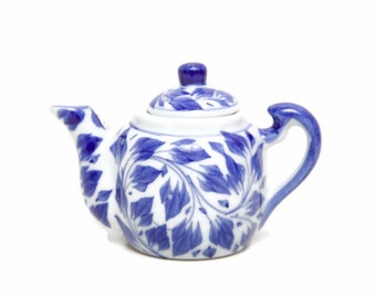 May include: A small, white ceramic teapot with a blue floral pattern. The teapot features a curved handle, a spout, and a lid with a small knob. The blue design includes leaf-like shapes. This teapot is suitable for brewing tea.