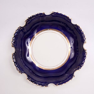 Könnte beinhalten: Eine dekorative Schale mit gewelltem Rand, mit einem weißen Zentrum und einem tiefblauen Rand. Der Rand ist mit goldenen Zierleisten versehen. Das Design der Schale ist symmetrisch und elegant, geeignet zum Servieren oder Ausstellen.