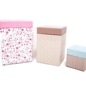May include: Three decorative gift boxes. The largest box has a pink lid and a white background with pink and tan floral patterns. The middle box has a tan lid and a tan background with white polka dots. The smallest box has a light blue lid and a striped base.