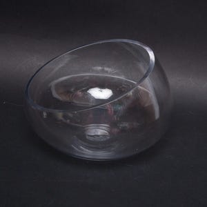 May include: A clear, slanted-rimmed glass bowl. The bowl has a rounded base and a smooth, transparent surface. The bowl is designed with a unique, asymmetrical shape, perfect for decorative displays or serving.