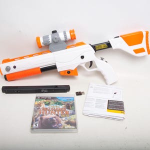 May include: A white and orange toy rifle with a scope, a black accessory, a game disc, a USB dongle, and a manual. The game disc is labeled "Cabela's Big Game Hunter 2012".