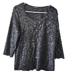 May include: Black long-sleeve top with a scoop neck and textured floral design. The top has a ruffled trim on the sleeves and a subtle striped pattern on the upper back. The top is hanging on a white hanger.