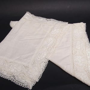 May include: Two off-white, folded handkerchiefs with intricate lace borders featuring floral patterns. The handkerchiefs are made of a soft, delicate fabric, and the lace adds a touch of elegance. The background is black.