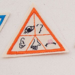 May include: A triangular patch with an orange border and a cream-colored interior. The patch is divided into four sections, each containing a simple black illustration: an eye, a question mark, an ear, a hand, and a hat.