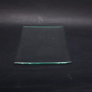 Puede incluir: Un plato cuadrado de vidrio con un borde ligeramente curvado. El plato es transparente con un sutil tinte verde a lo largo de los bordes. El plato está sobre un fondo oscuro.