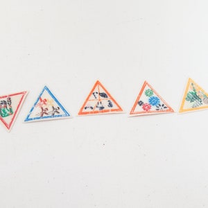 May include: Five triangular patches with various designs. The patches have a white background and are outlined in red, blue, orange, and yellow. The designs include floral patterns and abstract figures. These patches could be used for sewing or crafting projects.